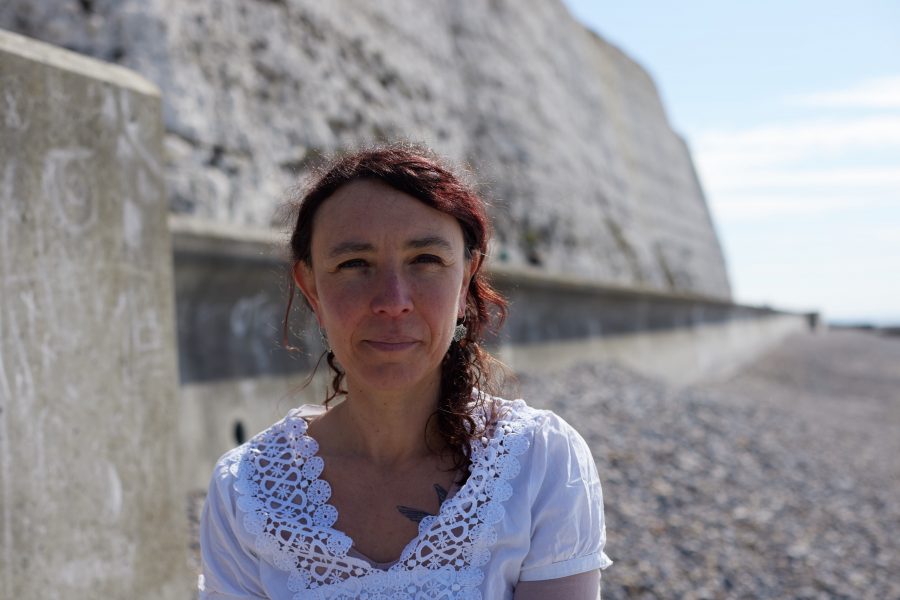 Adi Howarth standing in front of a chalk sea wall on the shingle, looking at the camera.